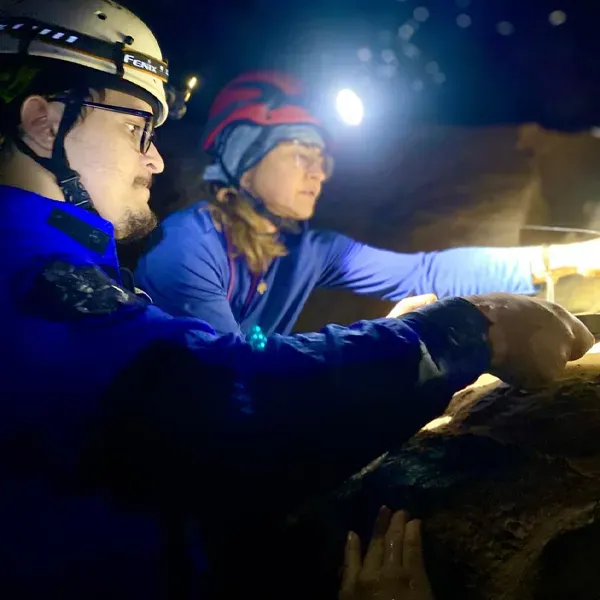 two people in a cave, each wearing a helmet with a light, they're looking at an animal just out of frame